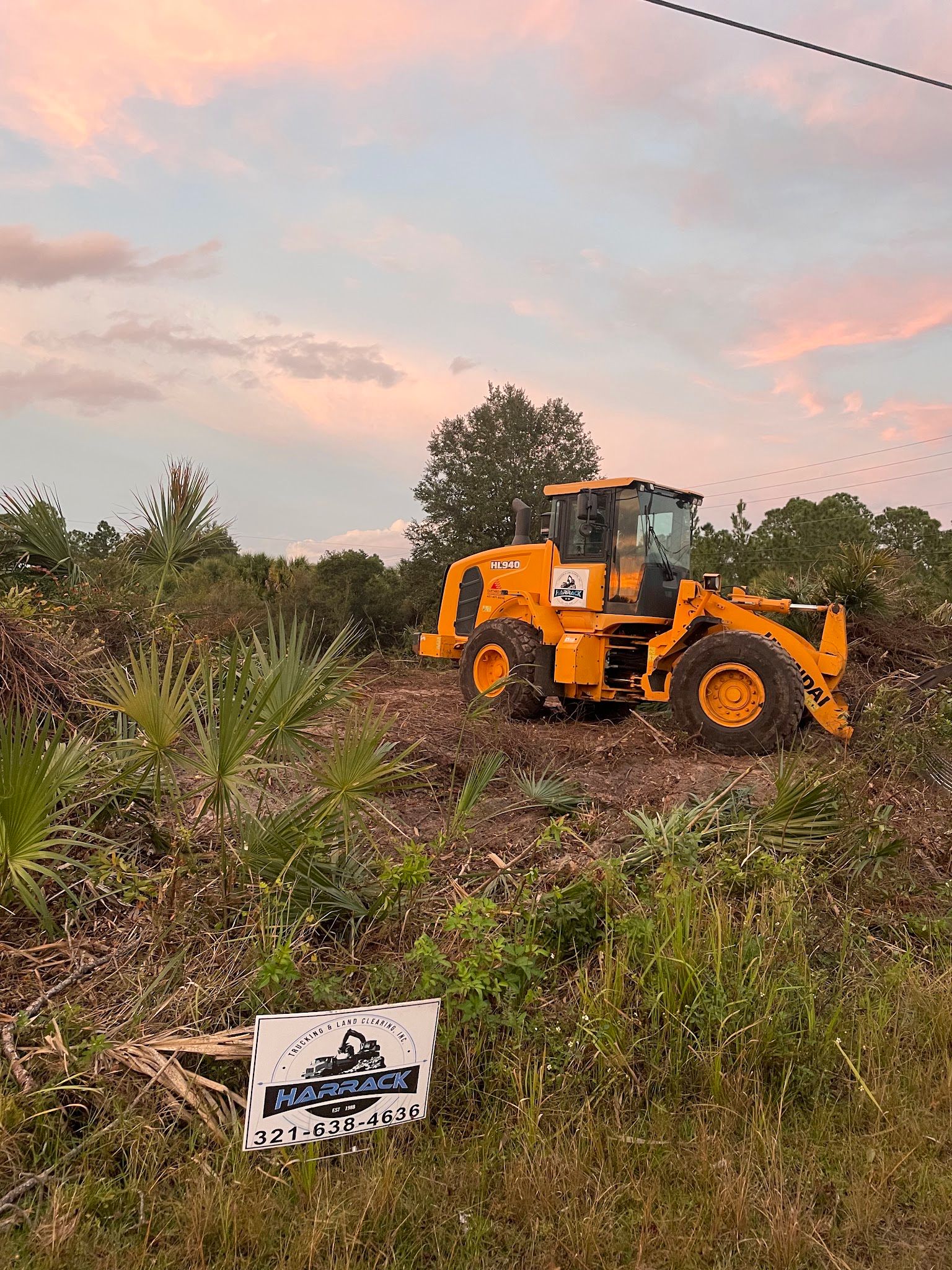 Yellow wheel loader clearing palmetto at sunset with a Harrack sign in front